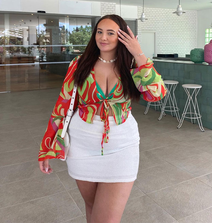 Influencer in a colorful outfit, standing indoors, smiling, with white stools and green decor in the background.