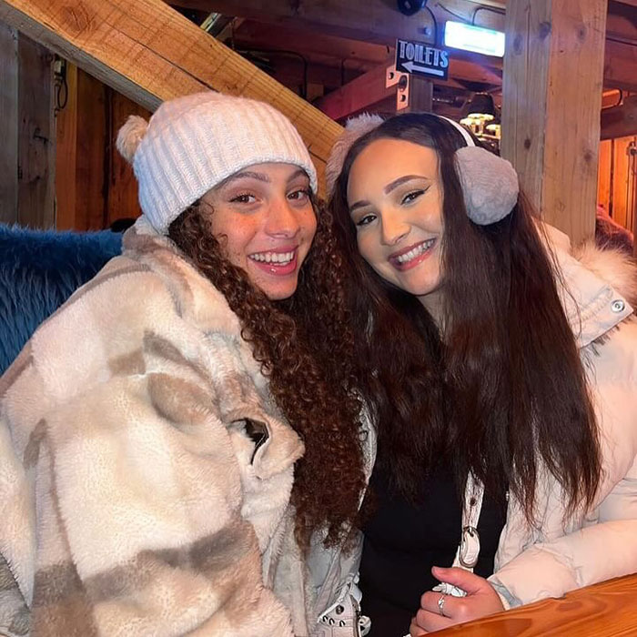 Two women smiling in warm coats and hats, at a cozy indoor location. Influencer trip.