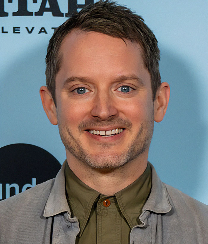 Man in a casual jacket smiling, standing in front of a blue background.