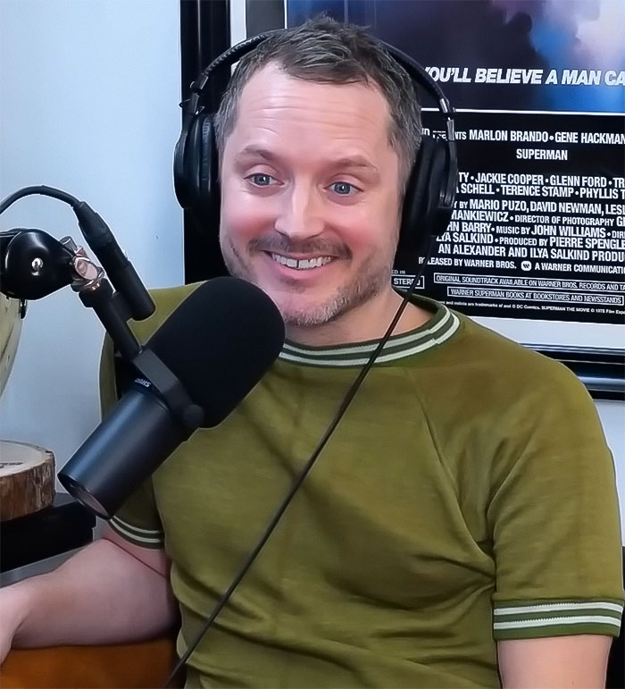 Elijah Wood smiling while speaking into a microphone, wearing headphones and a green shirt.