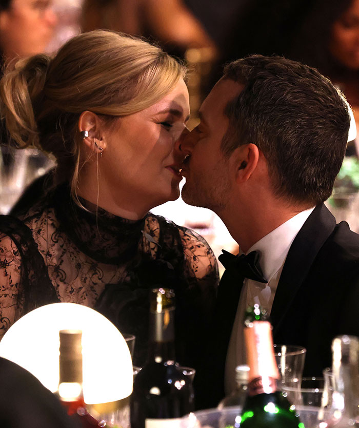 A couple shares a kiss at a formal event, surrounded by wine bottles and a glowing table lamp.