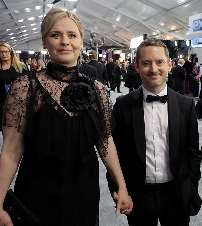 Couple holding hands on red carpet, both smiling, dressed elegantly in evening wear.