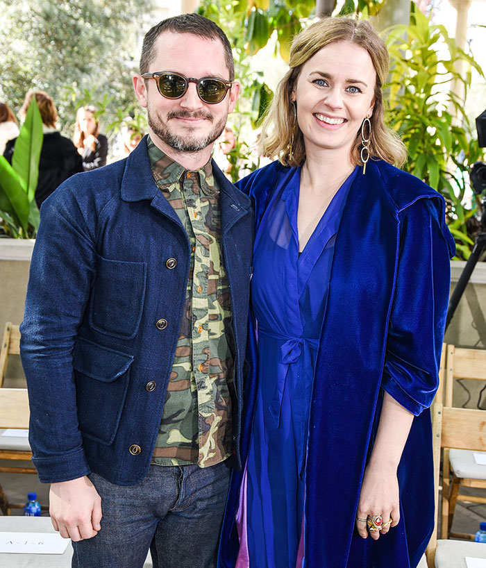 A couple dressed in casual and elegant attire, standing outdoors, surrounded by greenery and people.