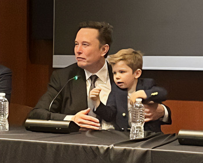 Man in a suit holding a child at a conference table with microphones and water bottles.