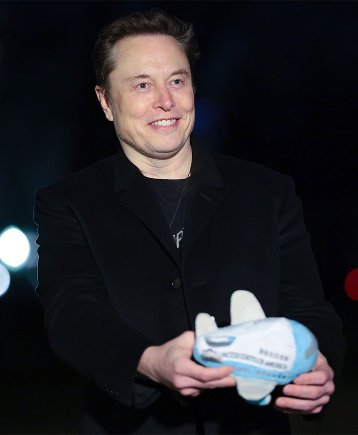 Elon Musk smiling while holding a toy rocket, wearing a black jacket at a public event.