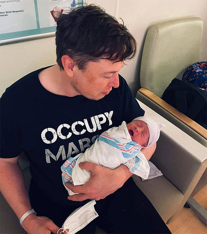 A person in a black shirt holds a newborn in a hospital setting, highlighting a family moment.