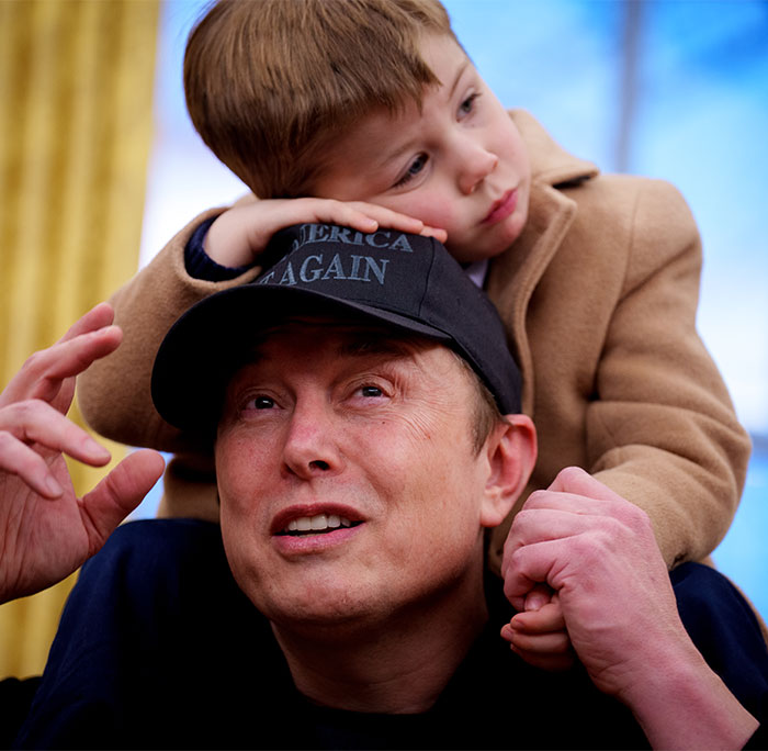 Elon Musk wearing a black cap with son X on his shoulders