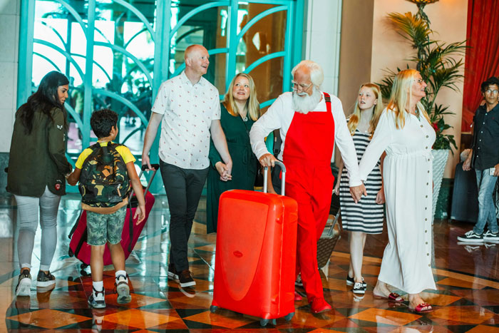 A childfree woman walks with a family at a hotel, a man in a white shirt and another in red overalls pulling a red suitcase.