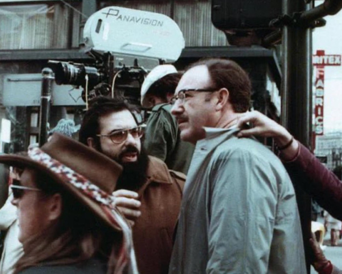 Film crew on set with cameras and equipment, featuring an actor in a jacket and glasses, showcasing a behind-the-scenes moment.