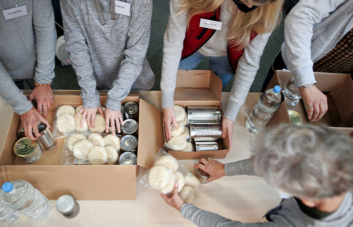 Volunteers packing food at a food bank, arranging canned goods and bread rolls in boxes.