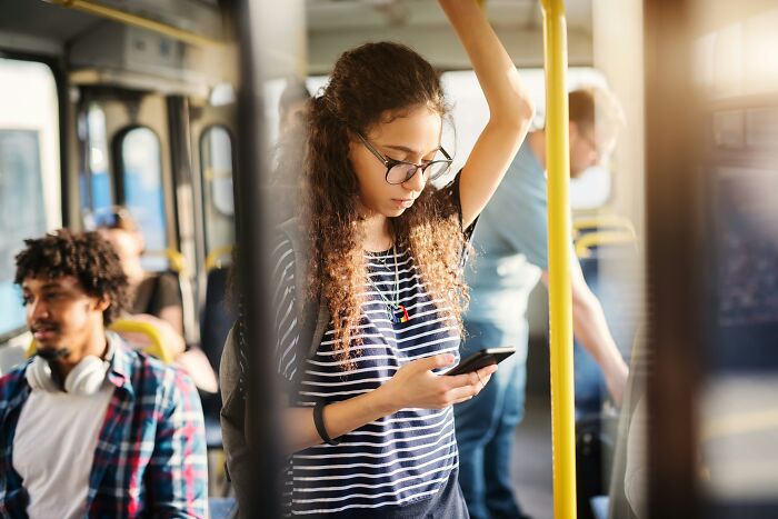 Gen Zer on a bus, wearing glasses and a striped shirt, focused on their phone.