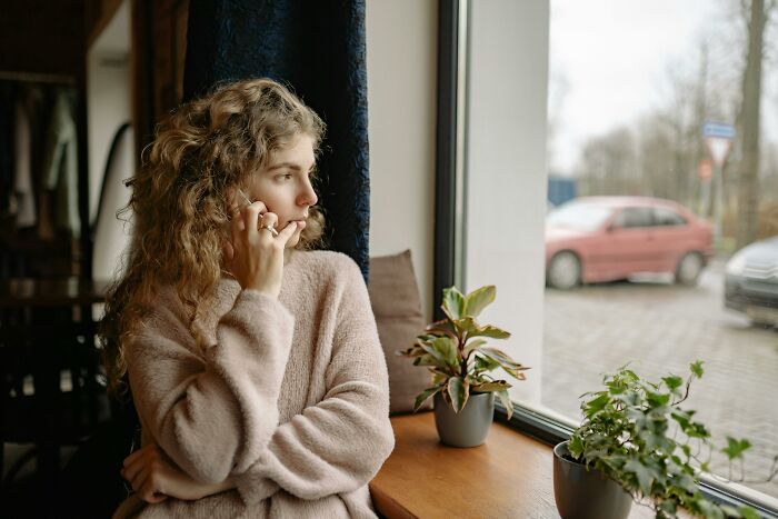 Young woman with curly hair gazing out window, surrounded by plants, reflecting a modern take on boomer complaints.