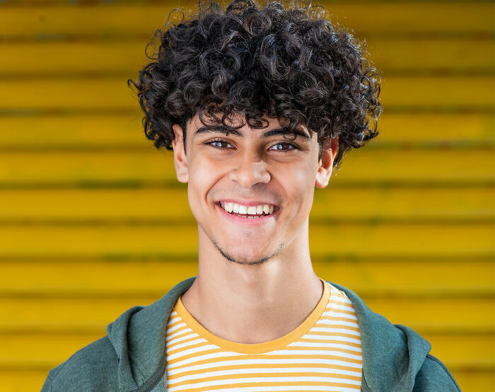 Gen Z person smiling, wearing a striped shirt and green jacket, stands against a yellow striped wall.