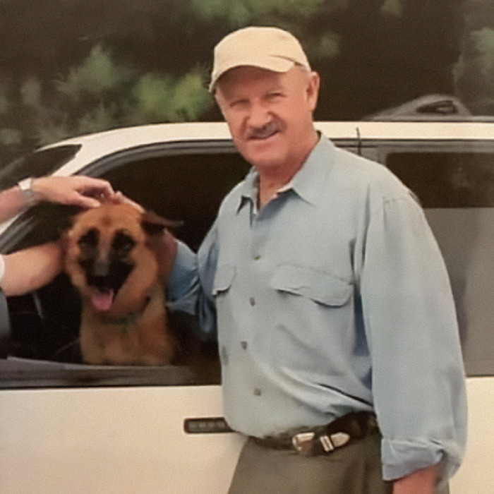 A man in a blue shirt and cap stands by a car with a dog inside, smiling outdoors.