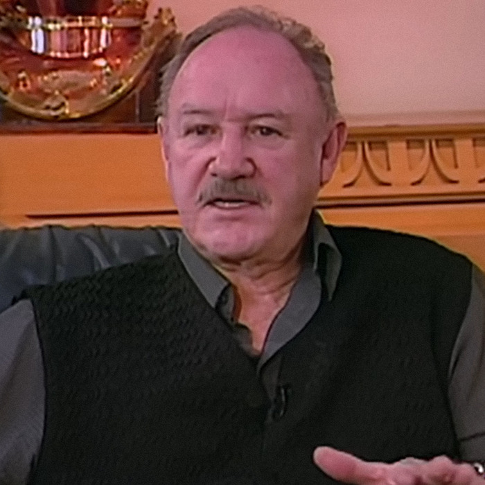 Gene Hackman in a black vest speaking at an interview.