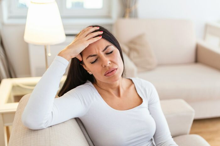 Woman in white sweater sitting on a couch with a hand on her forehead, looking concerned.