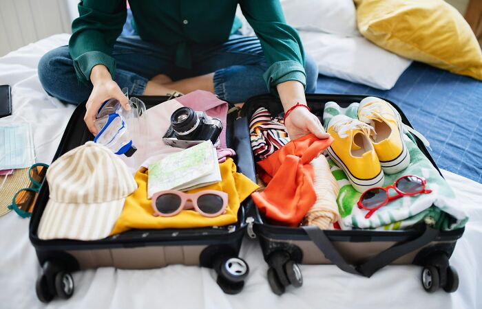 A person packing a suitcase with travel must-haves, including clothing, camera, shoes, and sunglasses, on a bed.