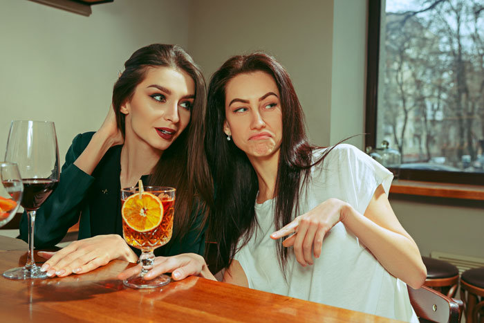 Two women at a bar having cocktails, one unimpressed, related to ex-girlfriend quarrel.