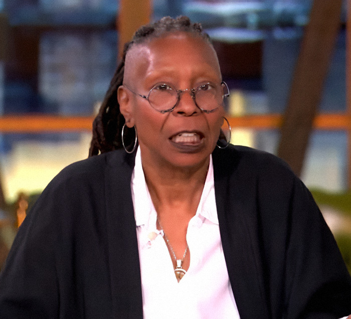 Woman wearing glasses and earrings speaking on a TV set.