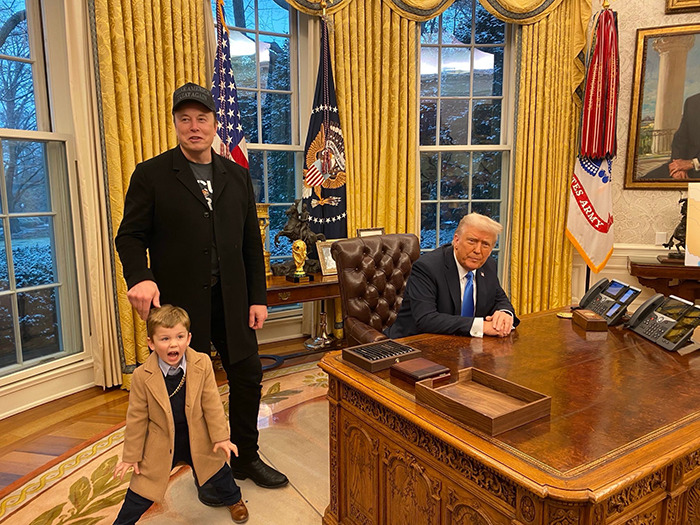 A man in a suit with a child in a coat standing next to someone seated at a large desk in an office with flags behind.