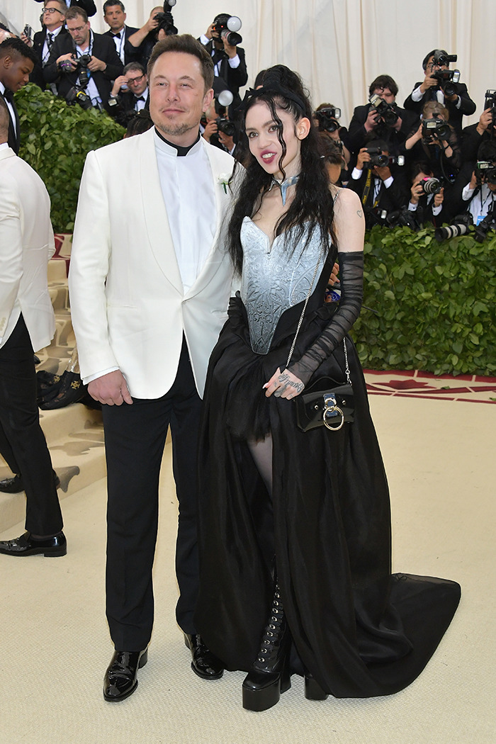 Grimes at a public event in a unique black and silver outfit with a white-clad companion, photographers in the background.