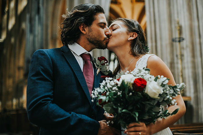 Couple kissing during wedding ceremony, holding a bouquet of red and white flowers.