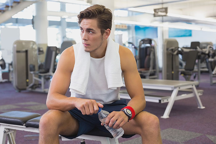 Man with towel in gym sitting, looking contemplative, holding a water bottle. Fitness equipment in background.