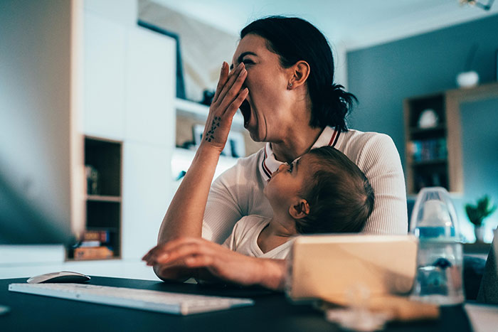 Woman yawning, holding baby, looking tired at desk; illustrating tiredness from new baby.