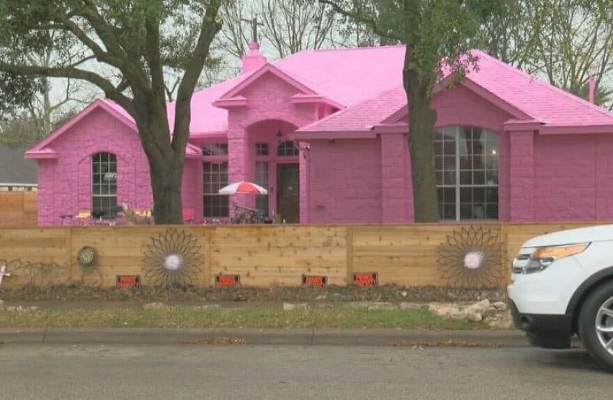 Pink house with mismatched architecture behind a wooden fence, surrounded by trees and a parked white SUV.