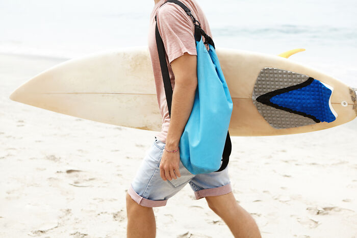 Person on beach carrying a surfboard and blue travel bag, illustrating packing essentials.