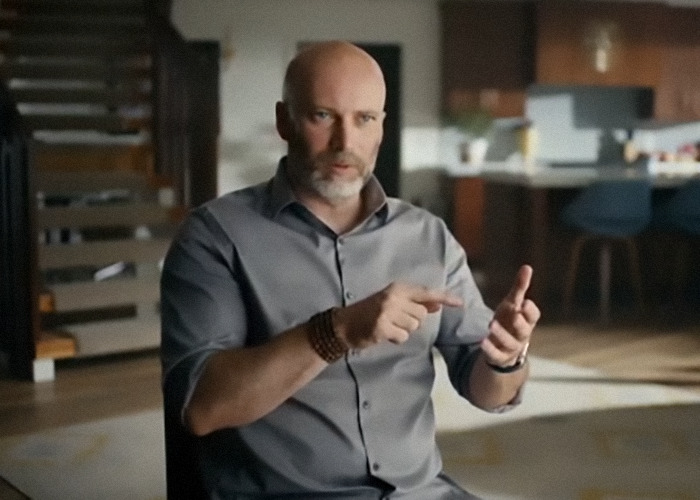 Man in an interview setting discusses momfluencer abuse, wearing a gray shirt and bracelet, in a modern living room.