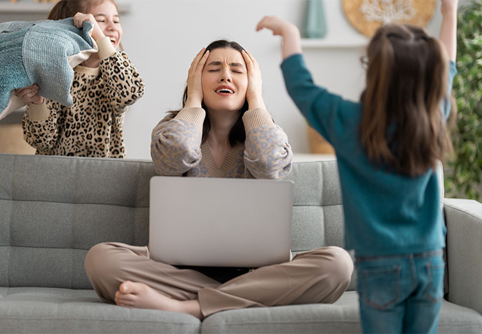 A stressed woman with a laptop on a couch as children play loudly around her, highlighting time management challenges at home.