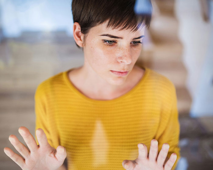 Woman in a yellow sweater, looking emotional, with her hands on a glass window.