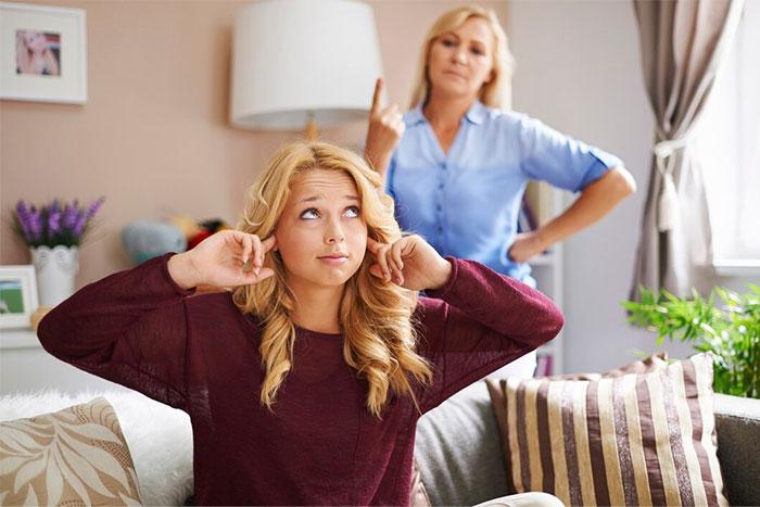 Mother scolding daughter covering ears in living room, expressing dislike for her name selected from Bible.