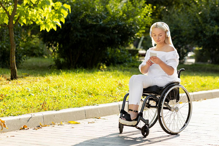 Woman in wheelchair outside, wearing headphones, reading a book, enjoying a sunny day with trees and grass around.