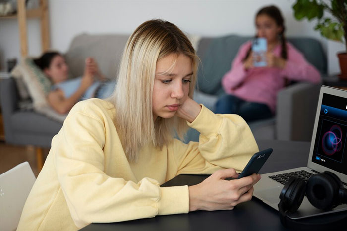 Teen girl looking at phone, appearing frustrated, while sitting near a laptop, wearing a yellow sweater in a living room.