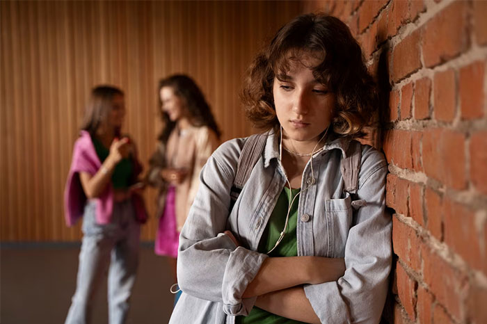 Teen girl upset by brick wall, classmates in background, dealing with name issues.