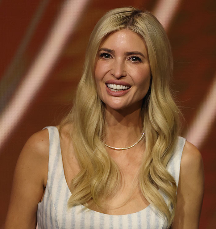 Blonde woman smiling with long hair extensions, wearing a striped dress and a necklace, in front of a warm background.