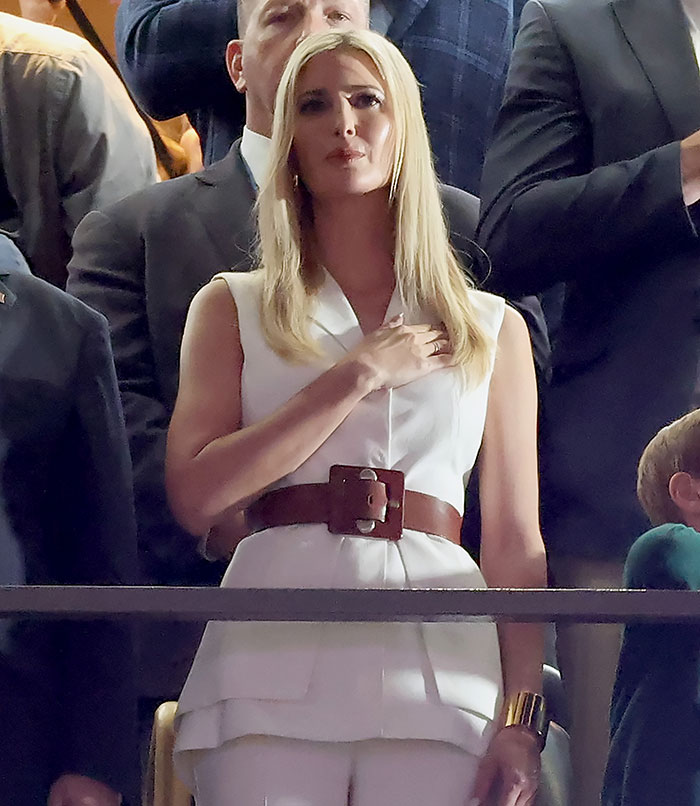 Blonde woman in a white outfit stands with hand on chest, surrounded by people, emphasizing hair extensions.