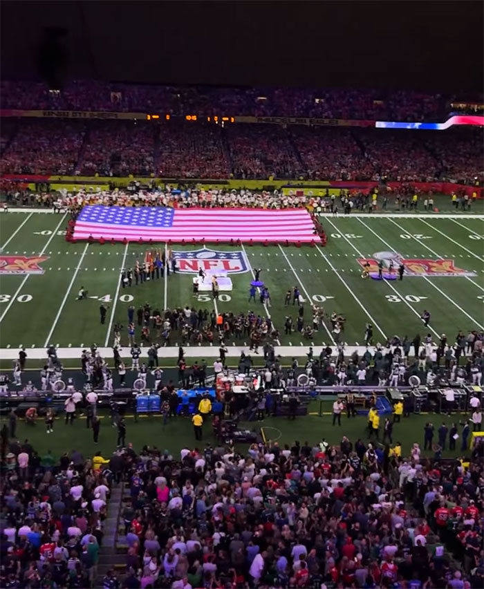 Super Bowl crowd with a large American flag on the field, pre-game ceremony in progress.