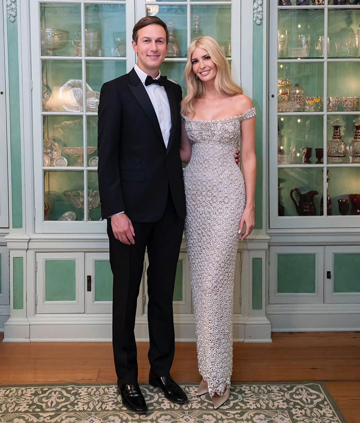 A couple in formal attire stands in front of a cabinet, with woman's hair styled using expensive extensions.