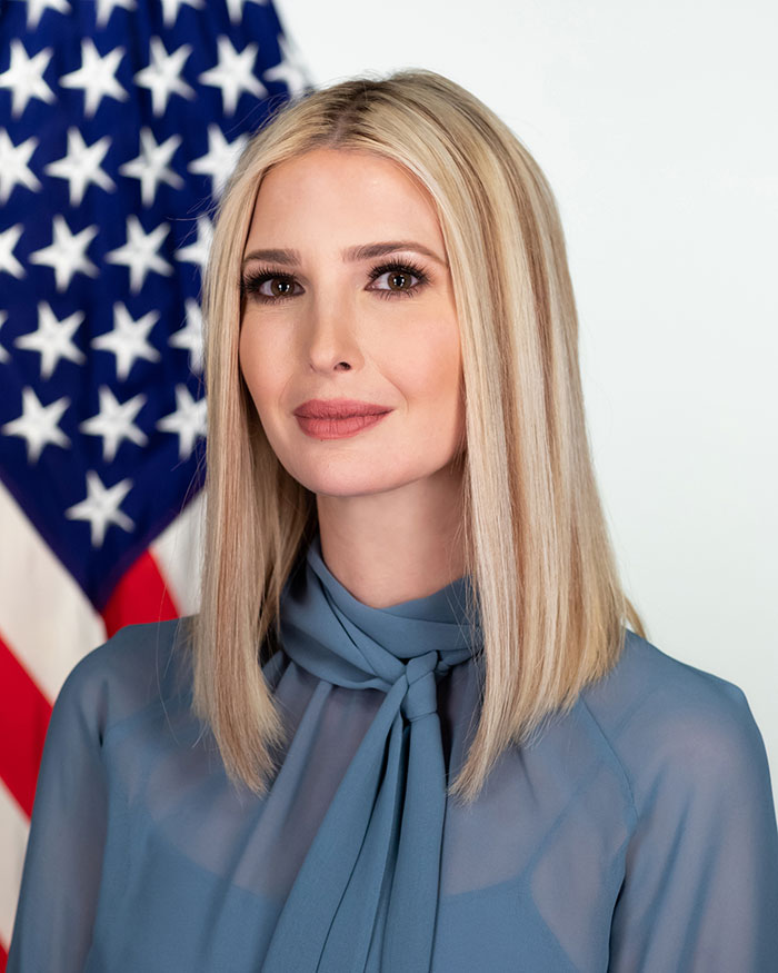 Woman with blonde hair extensions, wearing a blue blouse, stands in front of an American flag background.