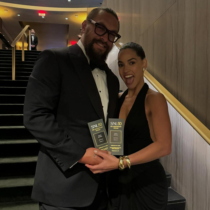 Jason Momoa on the red carpet with new girlfriend, both holding tickets and smiling at an event.