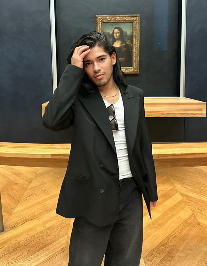 Celebrity hairstylist in a black suit poses in a gallery setting with a famous painting in the background.