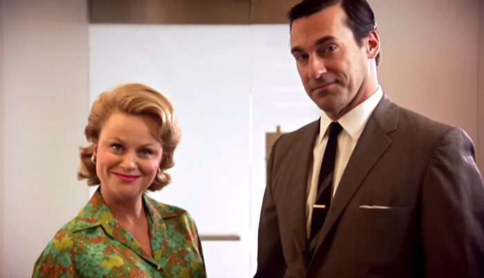 Two people in 1960s attire standing in an office setting, related to Jon Hamm's joke controversy.