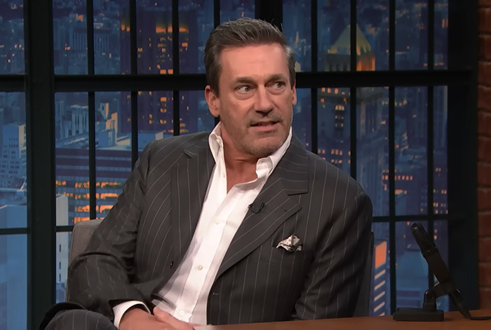 Man in a pinstripe suit on a talk show set, addressing controversy over Jon Hamm's joke involving Amy Poehler.