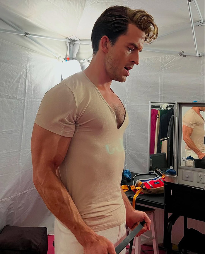 Jonathan Bailey in a fitted shirt holding a strap in a dressing room setting.