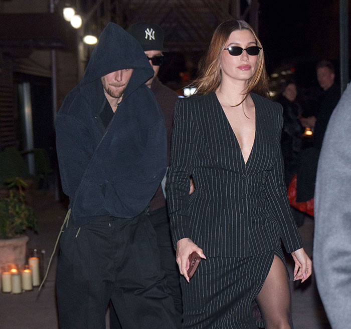 Two people walking; one in a hooded jacket, the other in a pinstripe dress, wearing sunglasses.