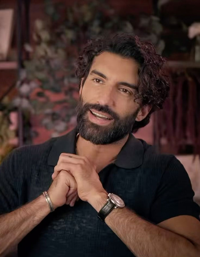 Young man with a beard, wearing a dark shirt and watch, smiling warmly in a cozy setting.