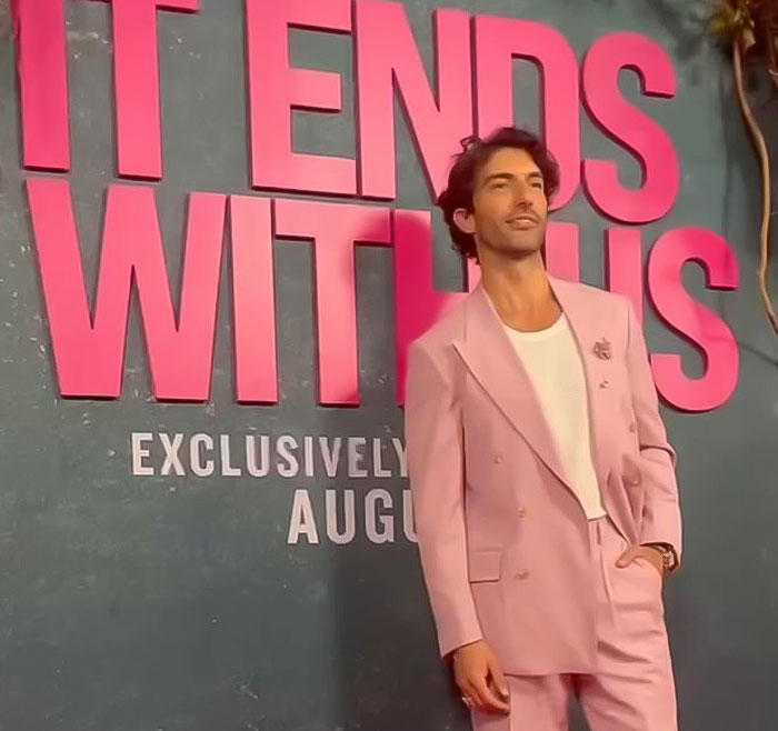 Man in a pink suit poses at "It Ends With Us" event, against a promotional background.